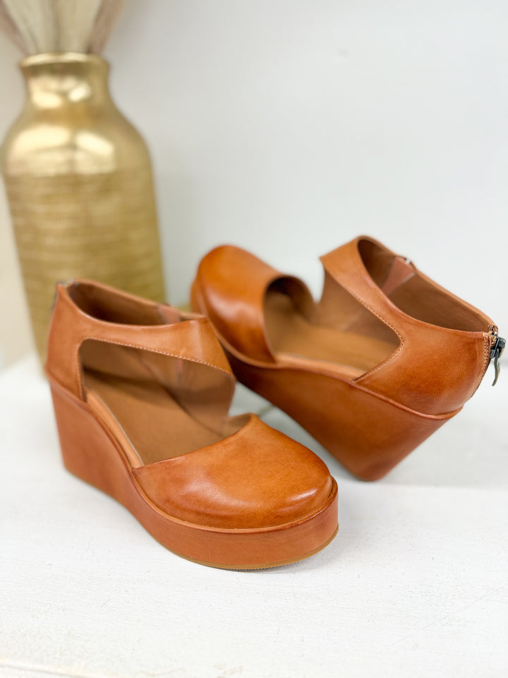 A pair of tan leather wedge shoes with cutout toes, displayed on a white surface with a gold vase in the background.