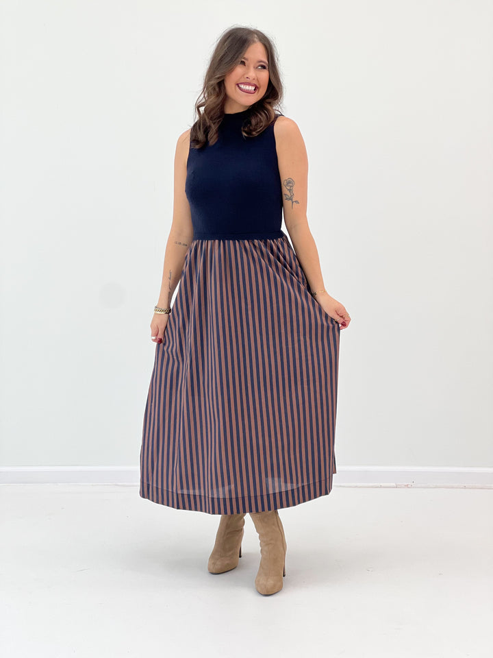 A woman modeling a sleeveless mock-neck midi dress with a navy blue top and a pleated skirt featuring vertical navy and rust-orange stripes, paired with beige ankle boots, against a plain white studio background.