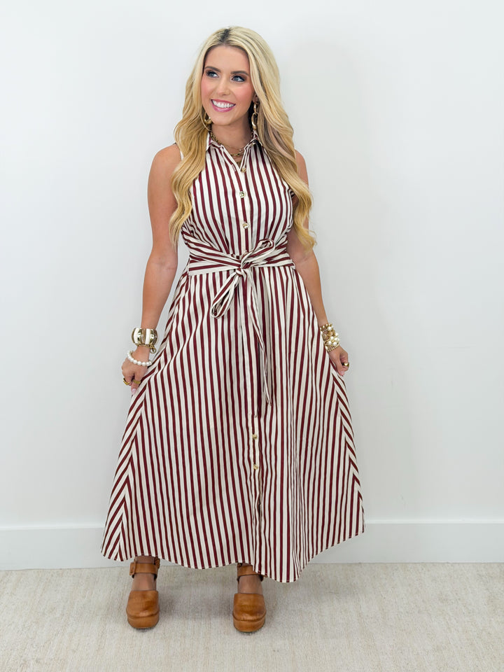A blonde woman models a sleeveless, high-neck maxi dress with vertical red and white stripes, featuring a tie-waist belt and button-down front. She is wearing brown platform clogs and gold bracelets, standing against a plain white wall.