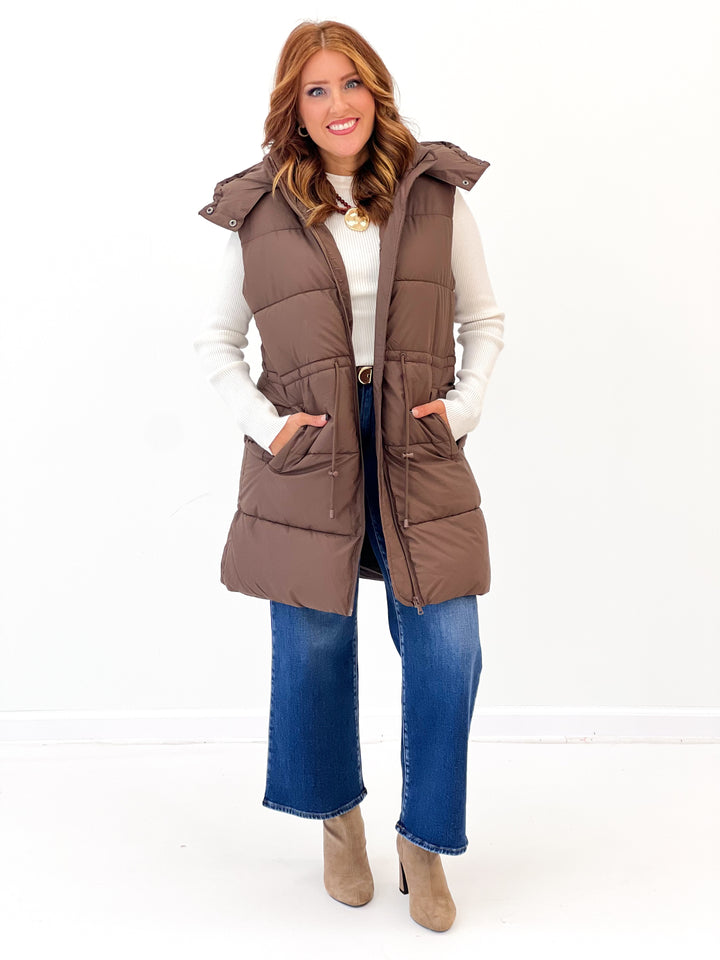 Brown longline quilted puffer vest with front zipper, belt loops, and side pockets