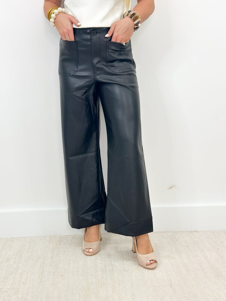 A person wearing black wide-leg faux leather pants with a white top and beige heeled sandals, standing against a plain white wall.