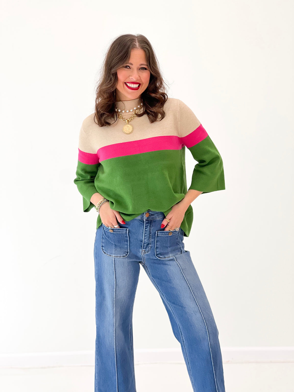 A woman modeling a color-block knit sweater with beige, pink, and green horizontal stripes, paired with blue jeans, against a plain white studio background.
