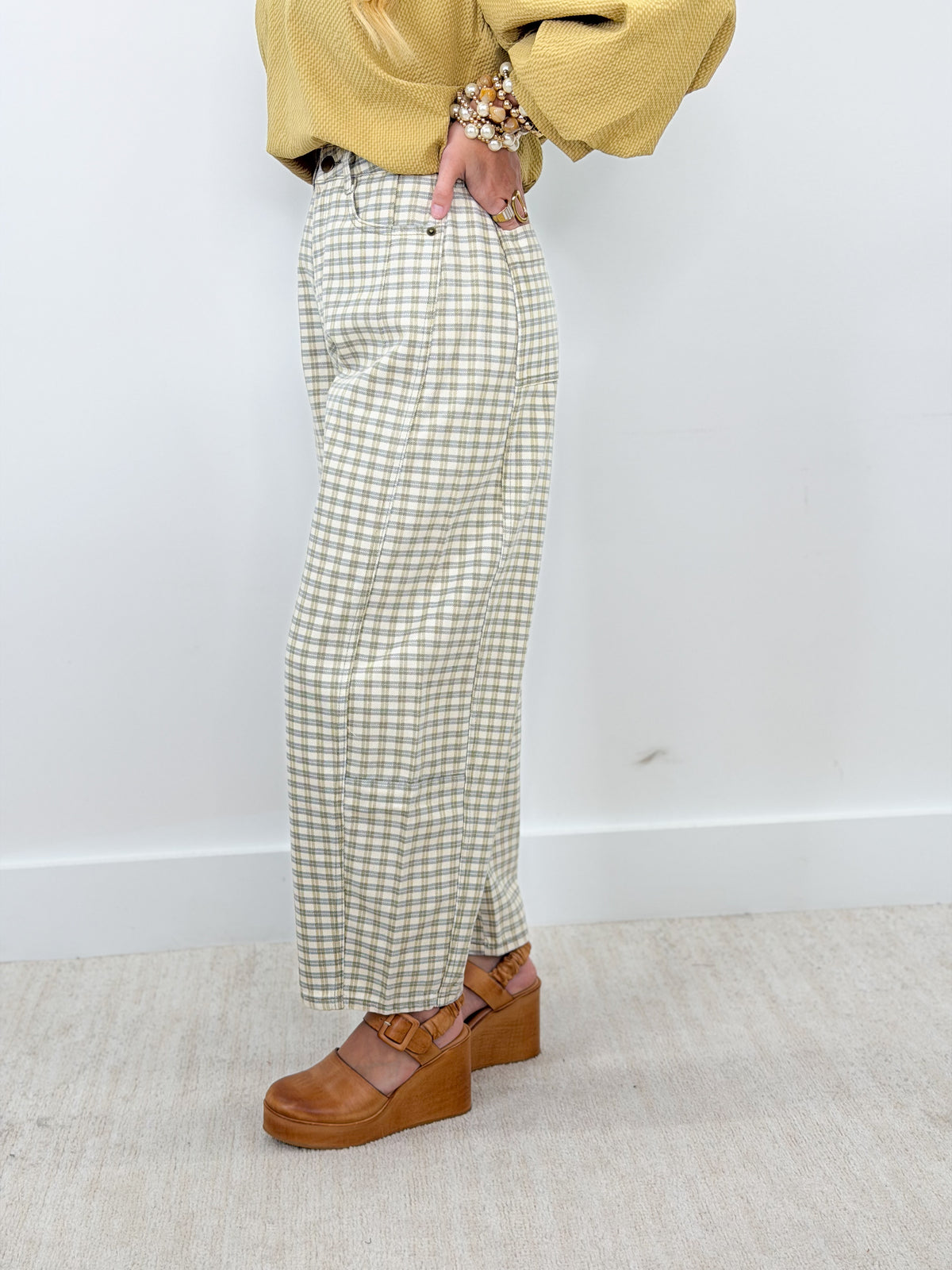 A person wearing high-waisted, wide-leg trousers in a beige and white gingham pattern, styled with a mustard yellow top and brown platform wedge sandals against a plain white wall and light beige floor.