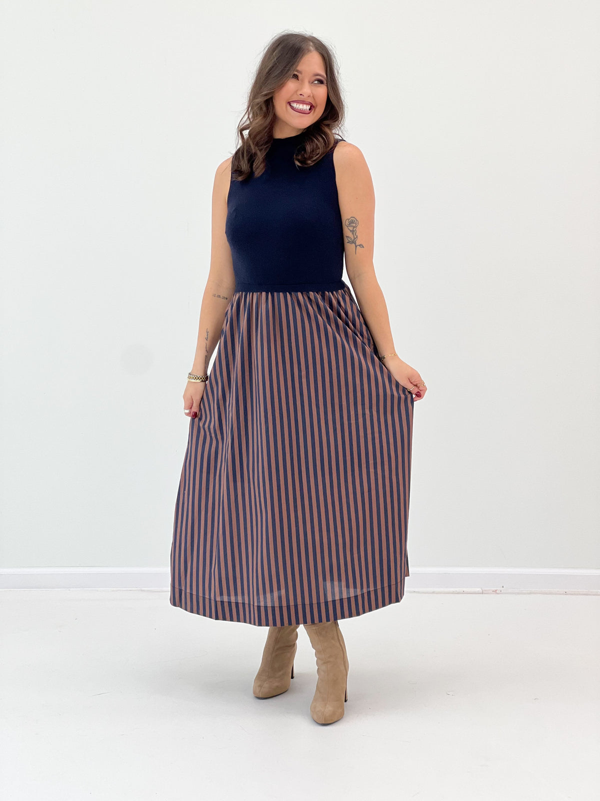 A woman modeling a sleeveless mock-neck midi dress with a navy blue top and a pleated skirt featuring vertical navy and rust-orange stripes, paired with beige ankle boots, against a plain white studio background.