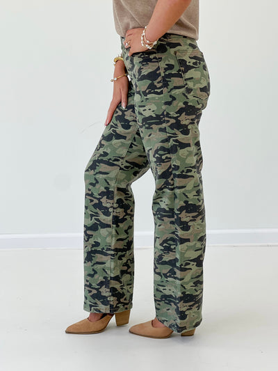 A person wearing camouflage wide-leg pants, styled with a beige top and tan pointed-toe heels, standing against a plain white studio background.
