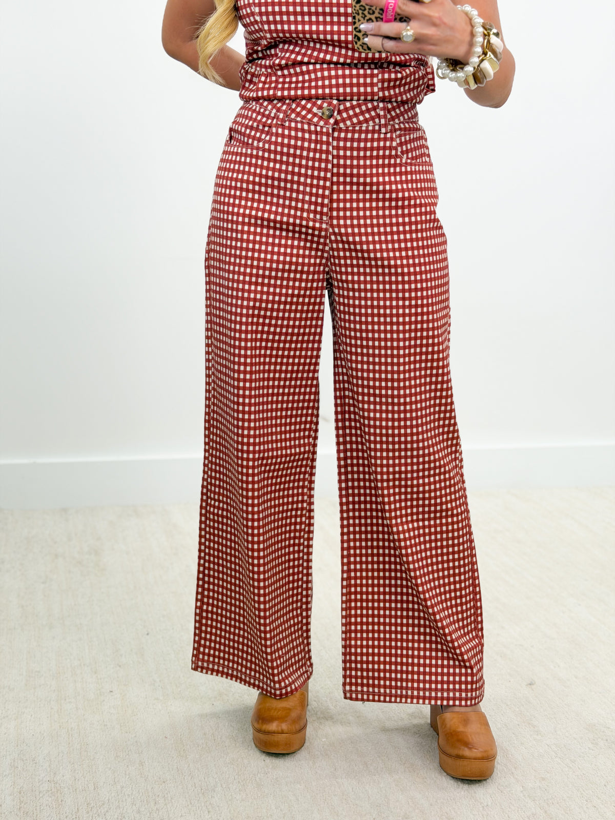 A person wearing red and white gingham wide-leg pants with a matching top, styled with brown platform shoes and gold jewelry, standing against a plain white wall.