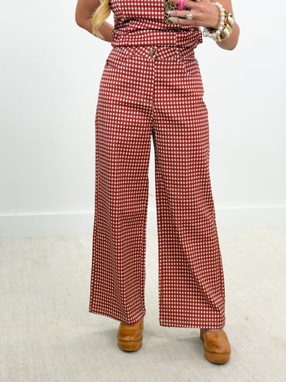 A person wearing red and white gingham wide-leg pants with a matching top, styled with brown platform shoes and gold jewelry, standing against a plain white wall.