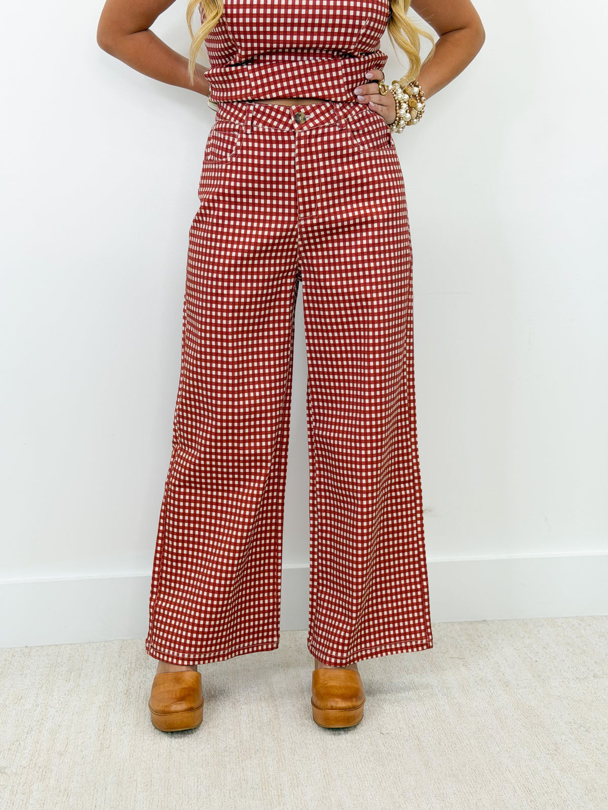A person wearing red and white gingham wide-leg pants with a matching top, styled with gold bracelets and brown platform shoes against a white wall and light carpet.
