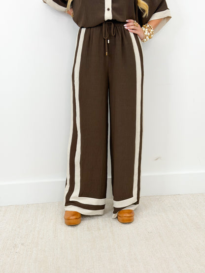 A person wearing brown wide-leg pants with thick cream side stripes and a drawstring waist, paired with a matching brown top and tan shoes, standing against a white wall.