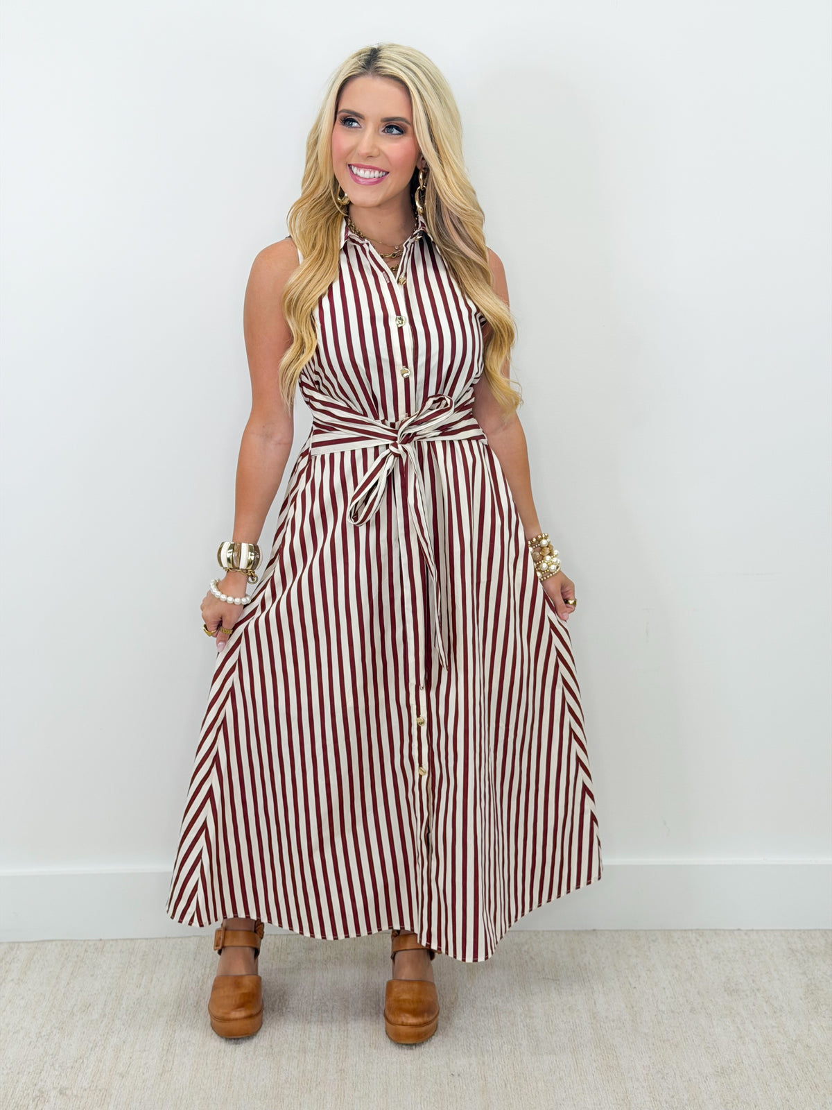 A blonde woman models a sleeveless, high-neck maxi dress with vertical red and white stripes, featuring a tie-waist belt and button-down front. She is wearing brown platform clogs and gold bracelets, standing against a plain white wall.