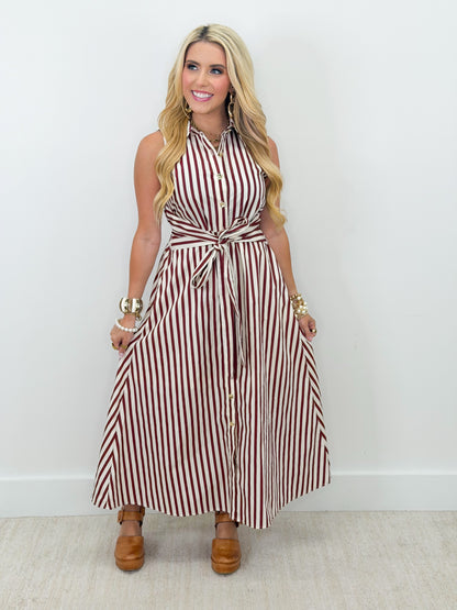 A blonde woman models a sleeveless, high-neck maxi dress with vertical red and white stripes, featuring a tie-waist belt and button-down front. She is wearing brown platform clogs and gold bracelets, standing against a plain white wall.