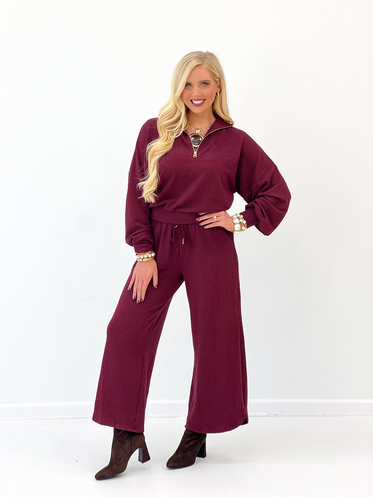 A woman modeling a matching burgundy two-piece lounge set consisting of a long-sleeve quarter-zip pullover and wide-leg pants, paired with brown heeled boots against a white studio background.