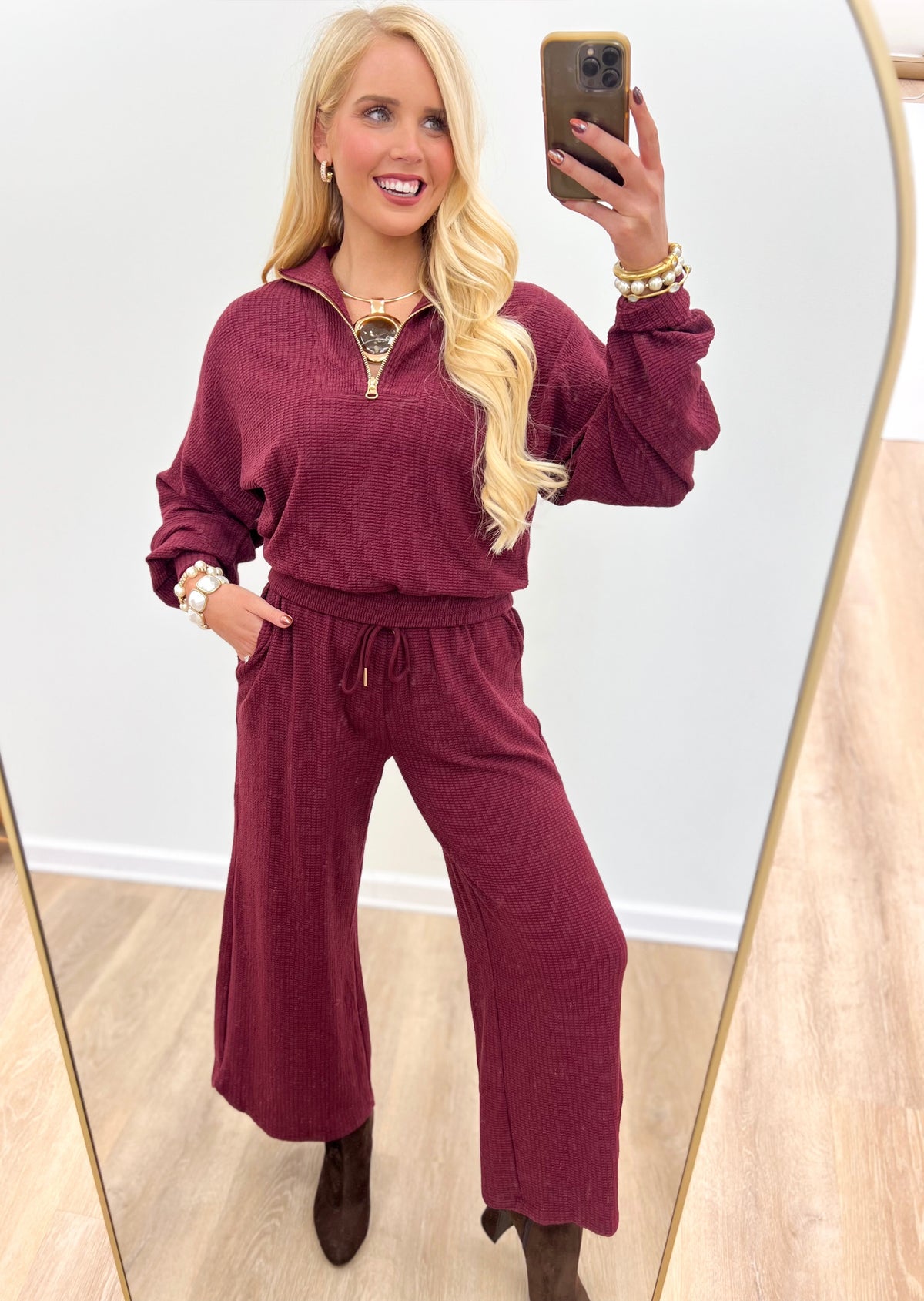 A woman takes a mirror selfie wearing a matching burgundy corduroy half-zip pullover and wide-leg pants set. She is standing in a room with light wood floors and a white wall, holding a gold smartphone.