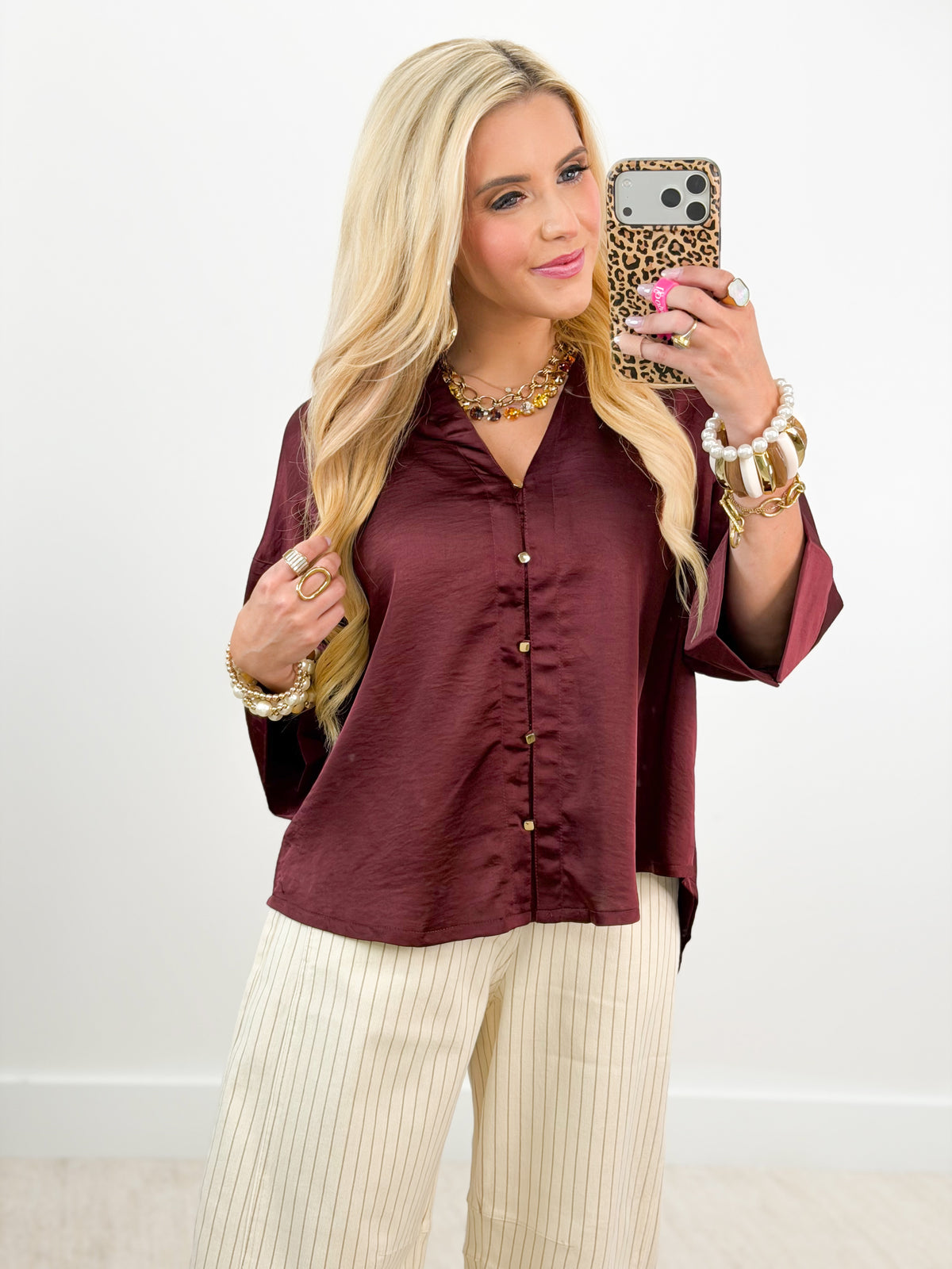 A blonde woman takes a mirror selfie wearing a long-sleeve, button-front satin blouse in a deep burgundy color, paired with cream-colored pleated pants. She is accessorized with gold bracelets, rings, and a statement necklace, holding a leopard-print phone case.