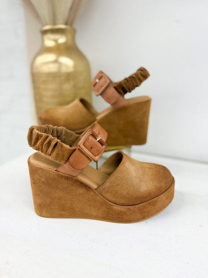 A pair of tan wedge sandals with ruched ankle straps and square buckle closures, displayed on a white surface with a blurred gold vase in the background.