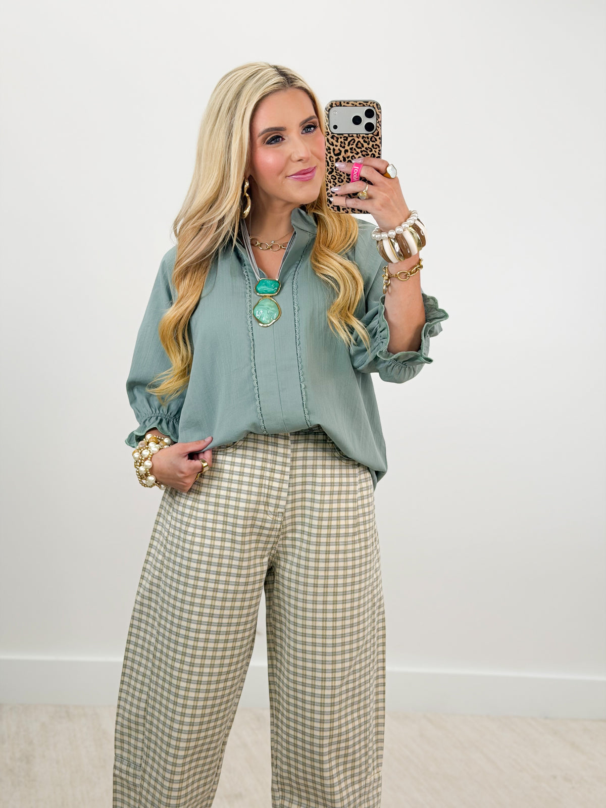 A woman takes a mirror selfie wearing a sage green long-sleeve blouse and beige-and-white gingham wide-leg trousers, accessorized with gold jewelry.