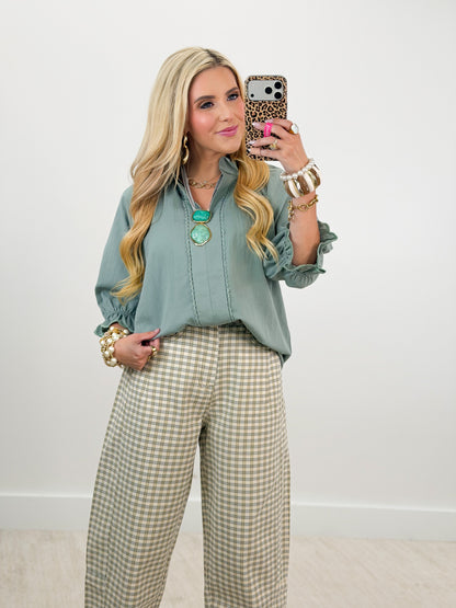 A woman takes a mirror selfie wearing a sage green long-sleeve blouse and beige-and-white gingham wide-leg trousers, accessorized with gold jewelry.