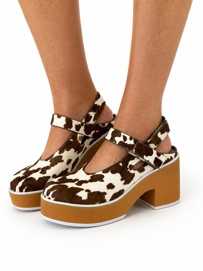 A pair of platform Mary Jane shoes with a brown and cream cow print upper, worn on a model's feet against a white background.