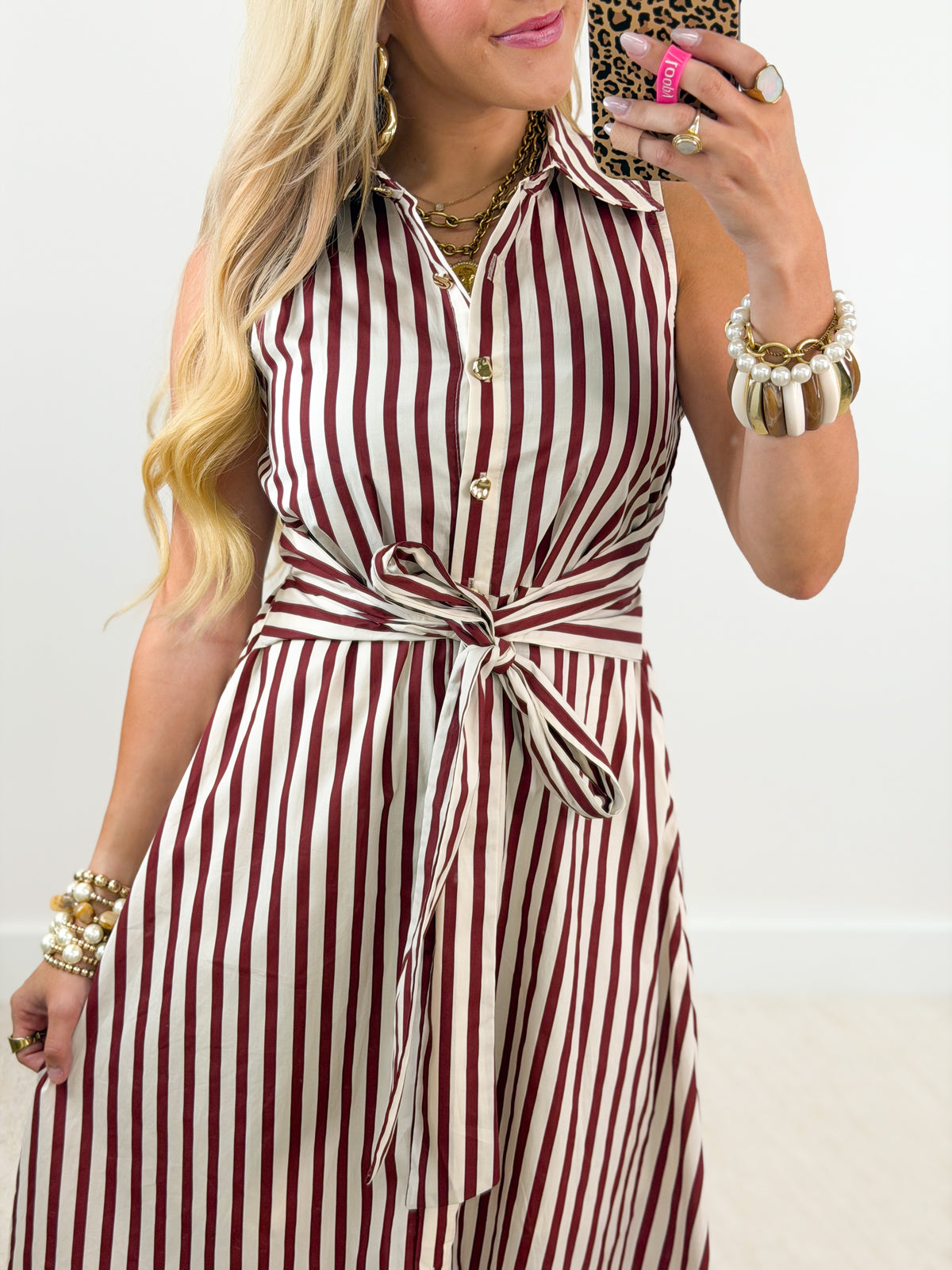 A woman wearing a sleeveless collared jumpsuit with vertical burgundy and cream stripes, holding a leopard-print phone in front of her face. She is accessorized with gold jewelry and bracelets against a plain white background.
