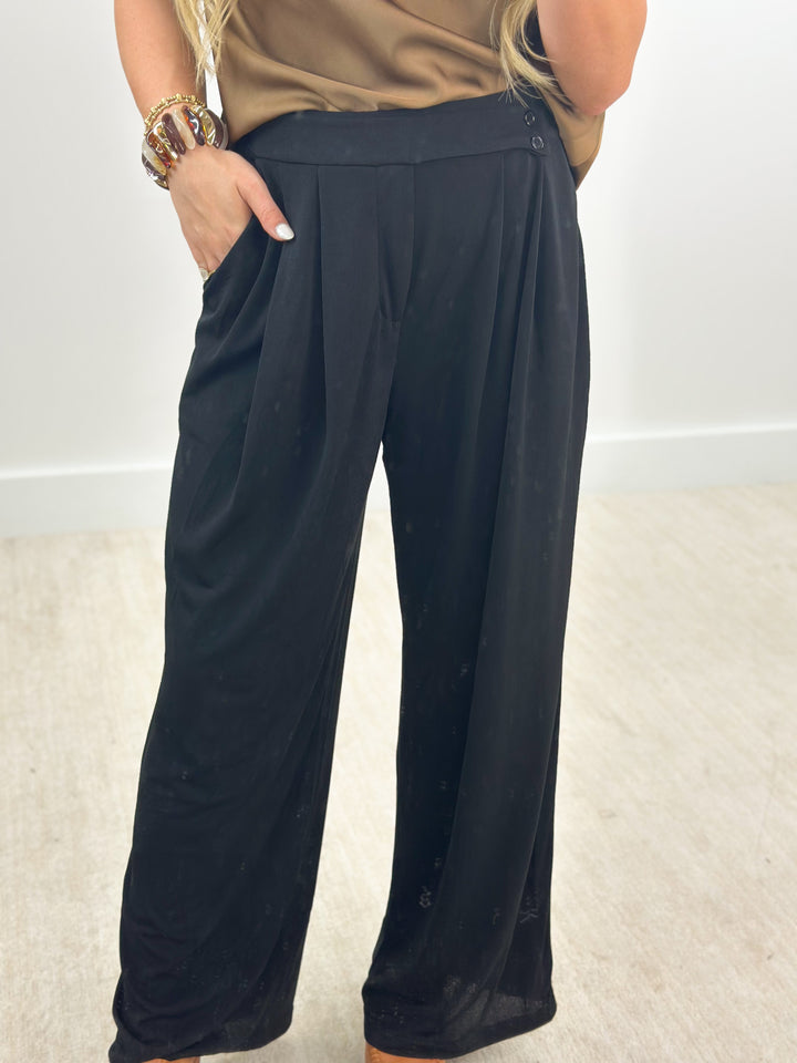 A person wearing loose black wide-leg pants with a tan top, standing indoors against a plain light wall and beige carpet.