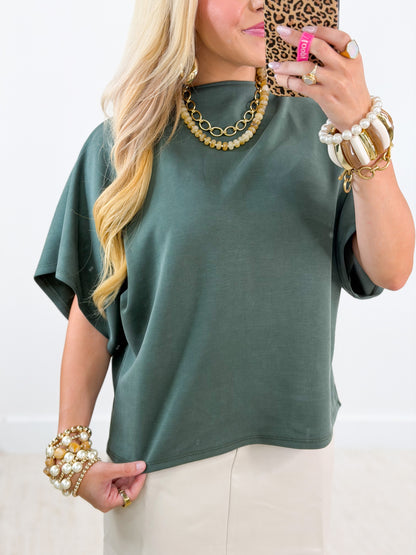 A woman takes a mirror selfie wearing a muted sage green short-sleeve top and cream pants, accessorized with gold necklaces, pearl and gold bracelets, and a leopard-print phone case.