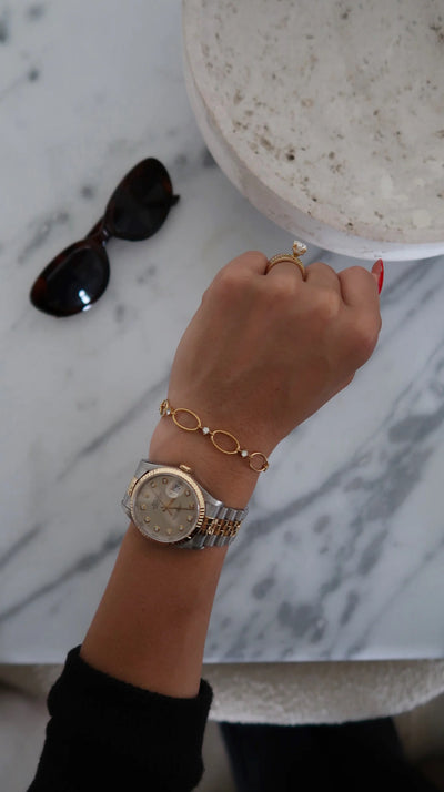Hand wearing a gold watch and bracelet on a marble surface with sunglasses.