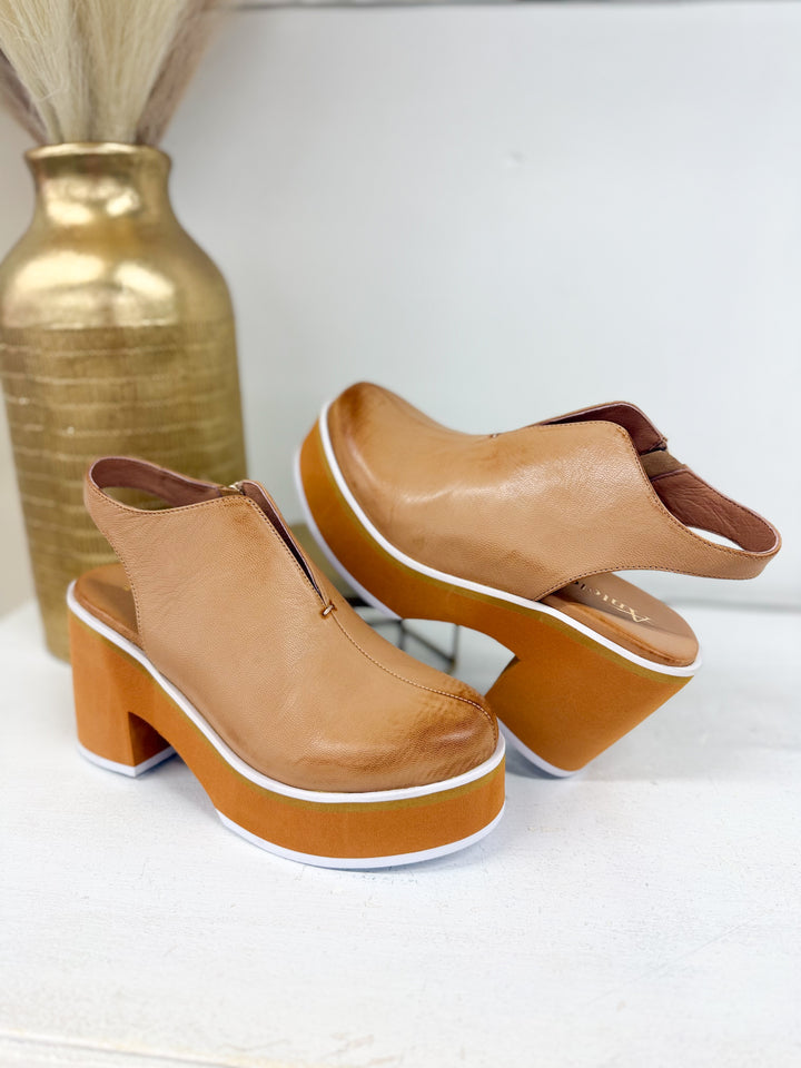 A pair of tan leather platform mule heels with chunky block heels and white trim, displayed on a white surface next to a gold vase.