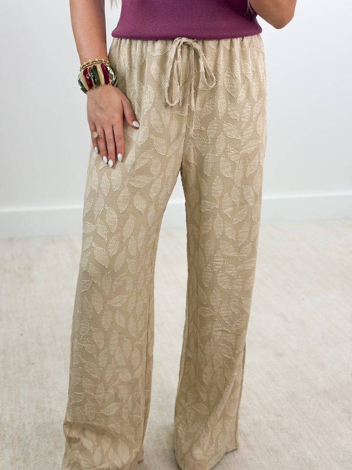 A person wearing beige wide-leg drawstring pants with a tonal leaf pattern, paired with a purple top. The photo is cropped from the waist down, showing the pants on a light-colored carpet against a white wall.