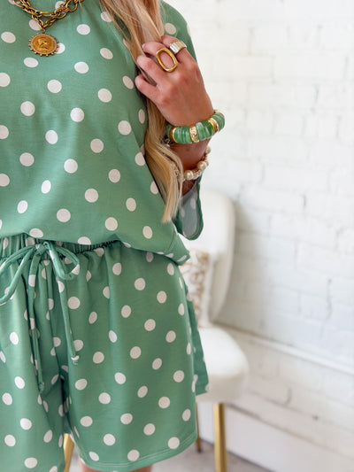 A person wearing a green and white polka dot romper, styled with gold jewelry, standing in front of a white brick wall.