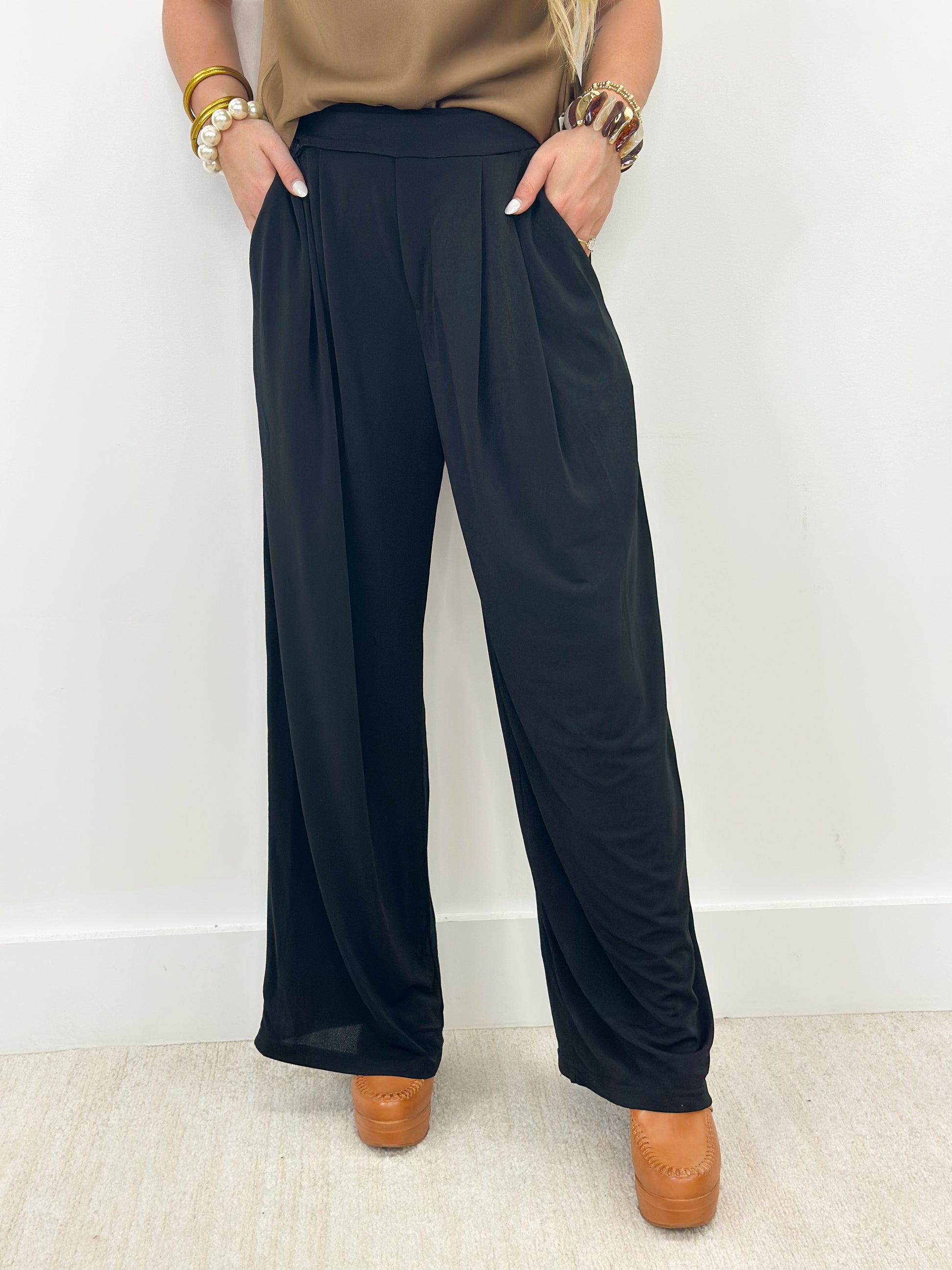 A person wearing black wide-leg pleated pants with a tan top and brown platform shoes, standing against a plain white wall.