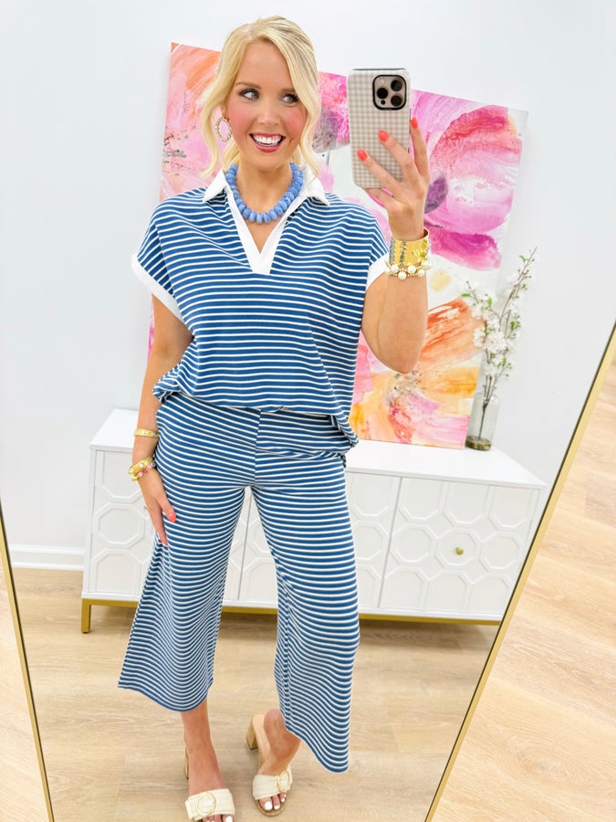 A woman takes a mirror selfie wearing a blue and white striped short-sleeve collared top with matching wide-leg cropped pants. She is wearing white heeled sandals, gold bracelets, and a chunky blue necklace in a brightly lit room with a white cabinet and colorful abstract wall art.