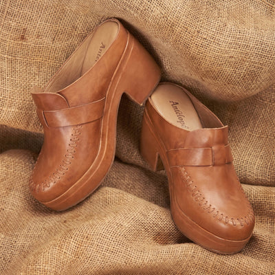 A pair of tan leather clogs with platform soles and stacked block heels, displayed on a textured burlap background. The brand name 'Autry' is visible on the insole.